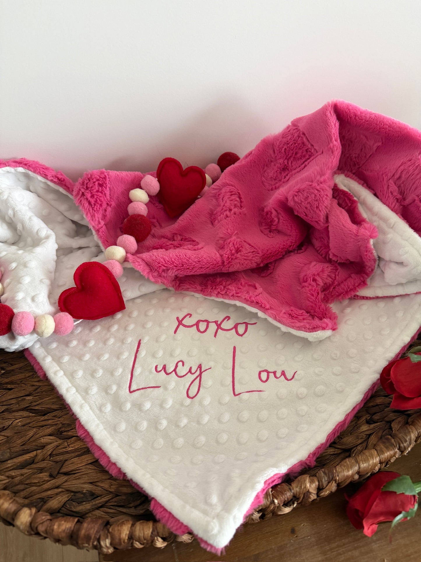 Baby blanket with pink trim and red hearts in a wicker basket on a wooden surface.