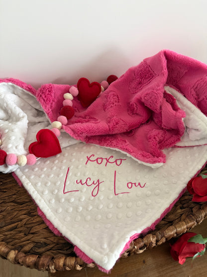 Baby blanket with pink trim and red hearts in a wicker basket on a wooden surface.