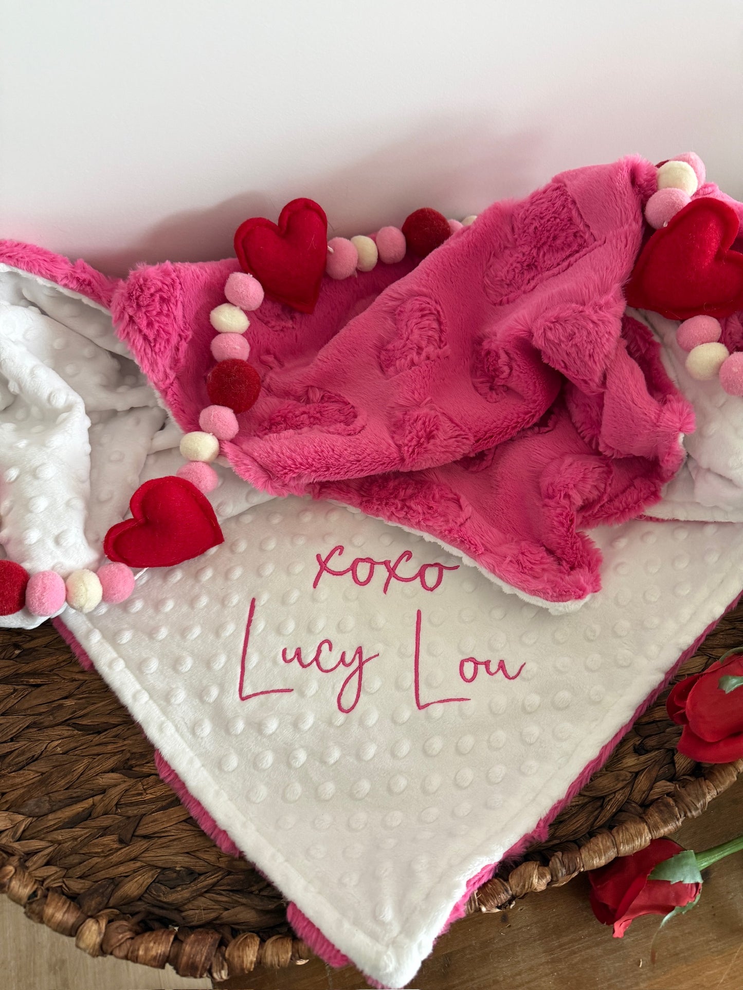 Pink and white baby blanket with 'xoxo Lucy Lou' text, surrounded by red hearts on a wooden surface.