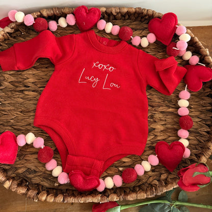Red baby onesie with 'xoxo Lucy Lou' text on a woven mat with heart decorations.