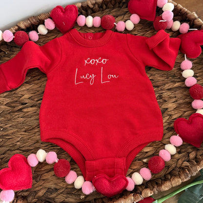 Red baby romper with 'XOXO Lucy Lou' text on a woven tray with heart decorations.