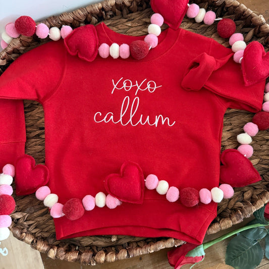 Red sweatshirt with 'XOXO Callum' text, surrounded by heart decorations on a woven basket.