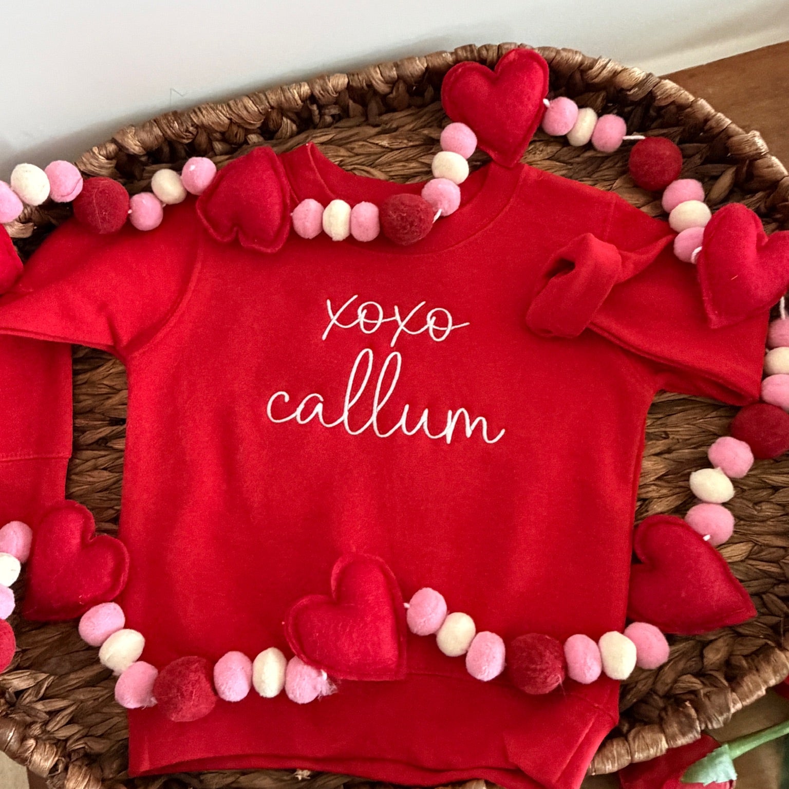 Red baby outfit with 'xoxo callum' text on a woven tray with decorative hearts and beads.