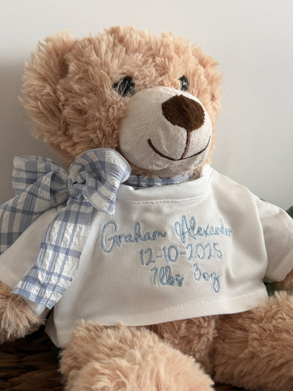 Teddy bear wearing a white shirt with embroidered text, sitting on a wooden surface.