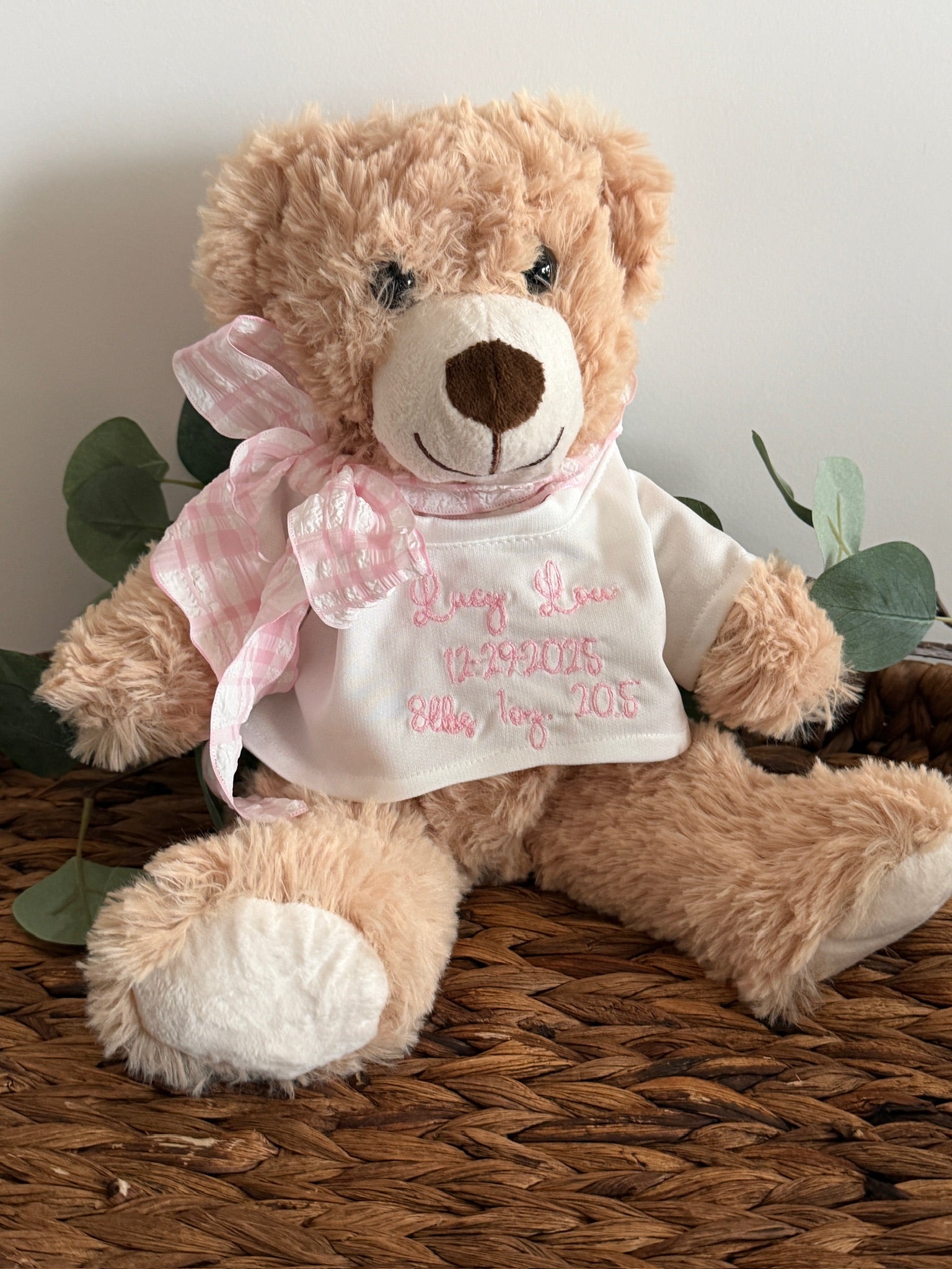 Teddy bear wearing a white shirt with pink text, sitting on a woven surface with green leaves.