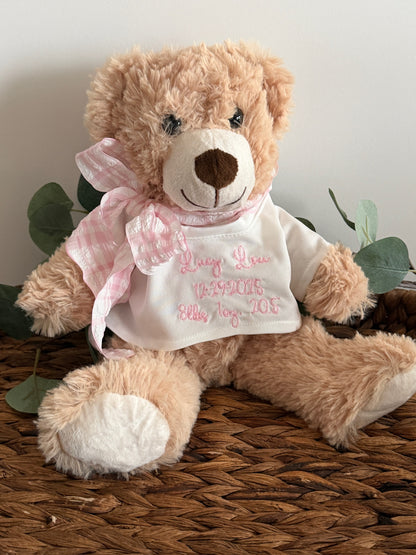 Teddy bear wearing a white shirt with pink text, sitting on a woven surface with green leaves.