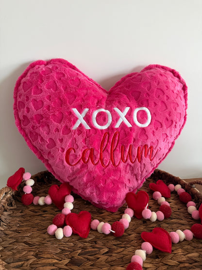 Heart-shaped pillow with 'XOXO callum' text on a woven surface with decorative hearts and beads.