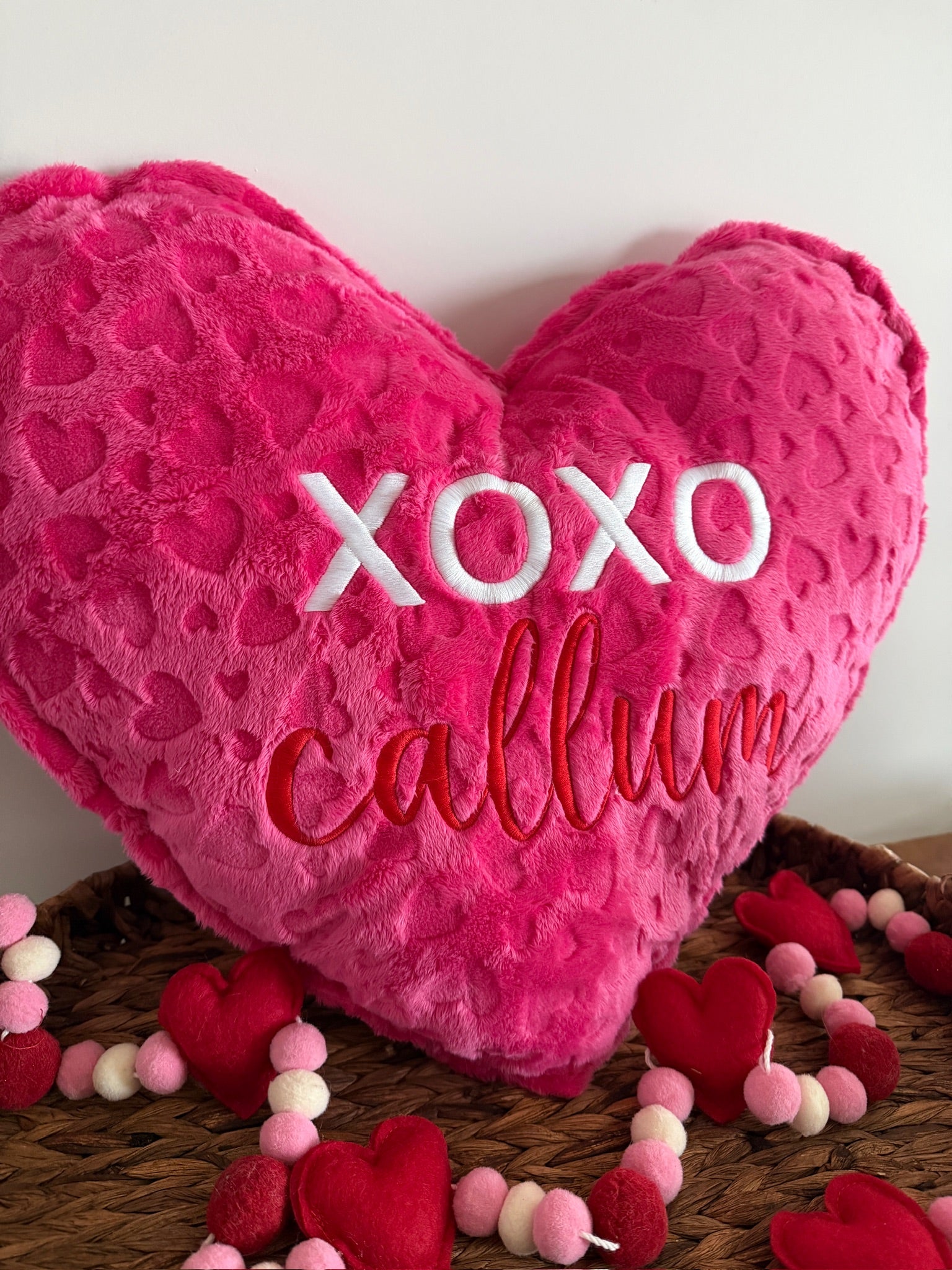 Heart-shaped pink pillow with 'XOXO callum' text on a wooden surface with small heart decorations.