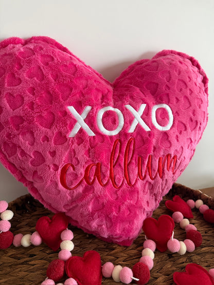 Heart-shaped pink pillow with 'XOXO callum' text on a wooden surface with small heart decorations.
