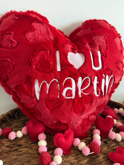 Red heart-shaped pillow with 'I §U martin' text on a wooden surface with decorative hearts.
