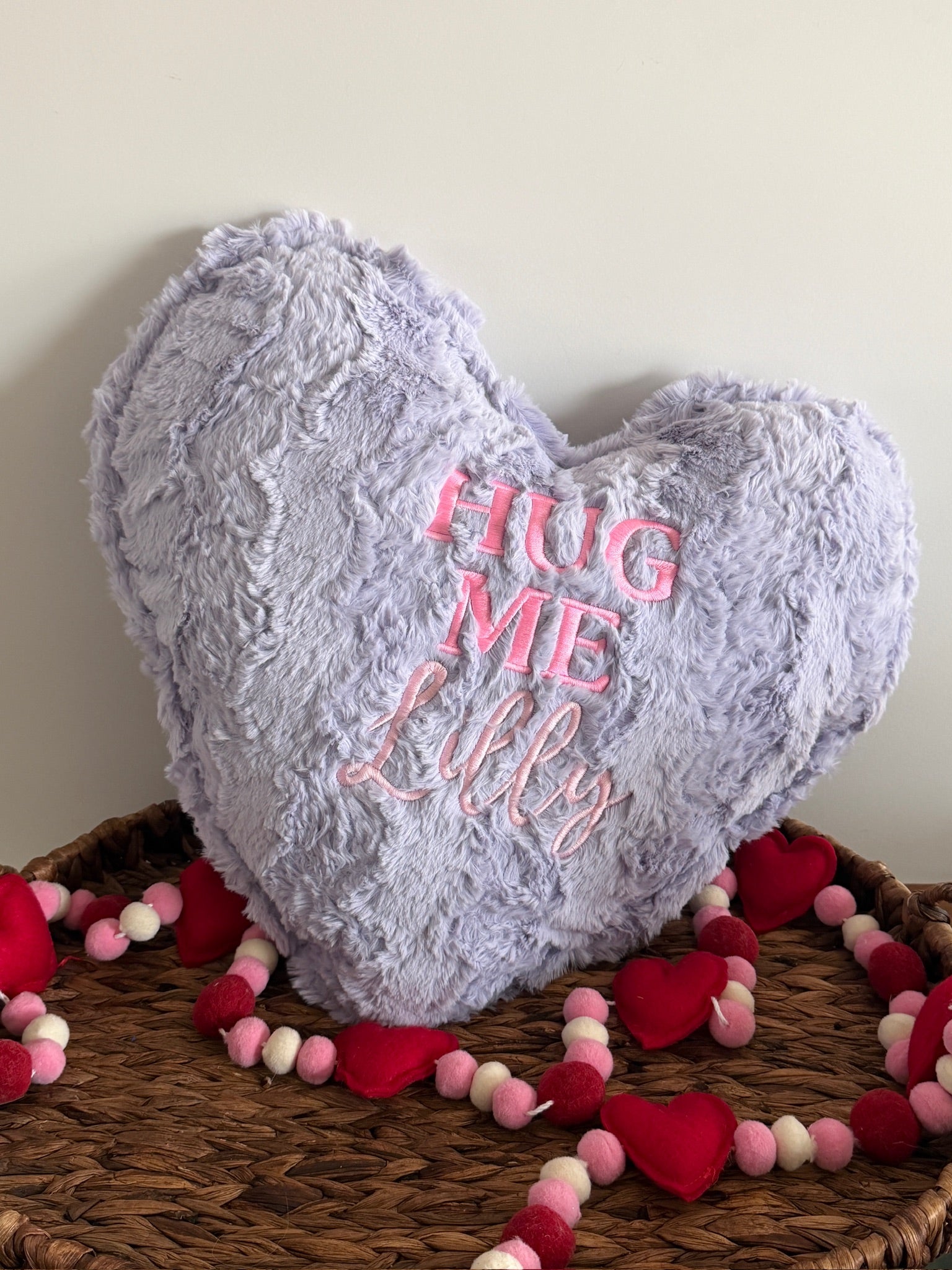 Heart-shaped plush pillow with 'Hug Me Lilly' text on a woven surface with decorative hearts and beads.