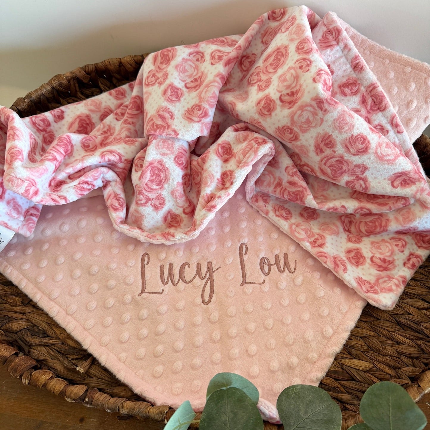 Pink baby blanket with red floral pattern and 'Lucy Lou' embroidery, placed in a woven basket.