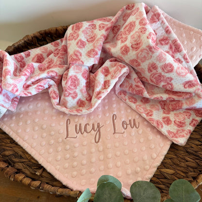 Pink baby blanket with red floral pattern and 'Lucy Lou' embroidery, placed in a woven basket.