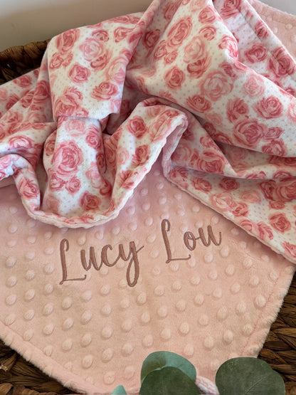 Floral patterned blanket with 'Lucy Lou' embroidery on a pink textured fabric background.