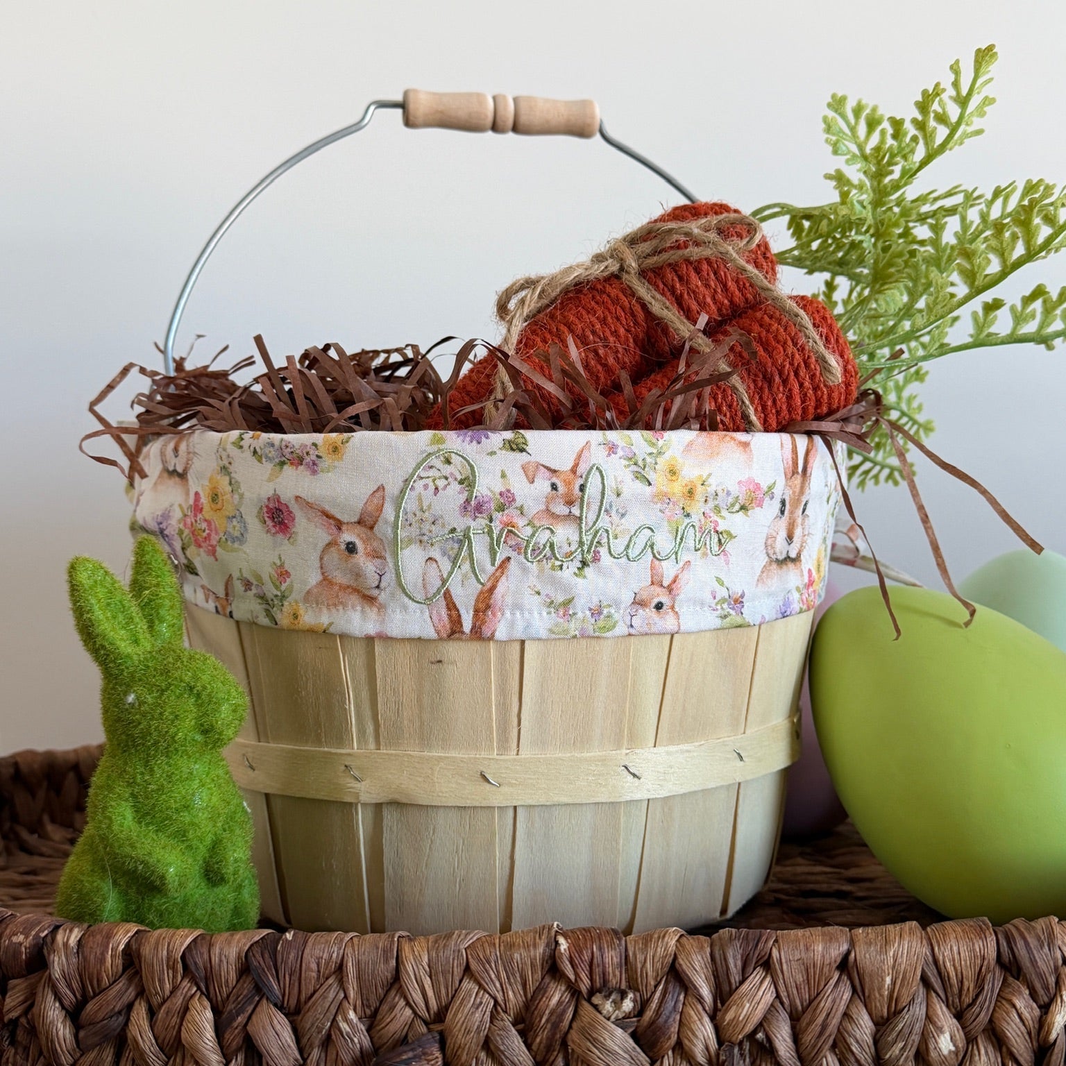 Decorative Easter basket with floral pattern, carrots, and a green bunny toy on a wooden surface.