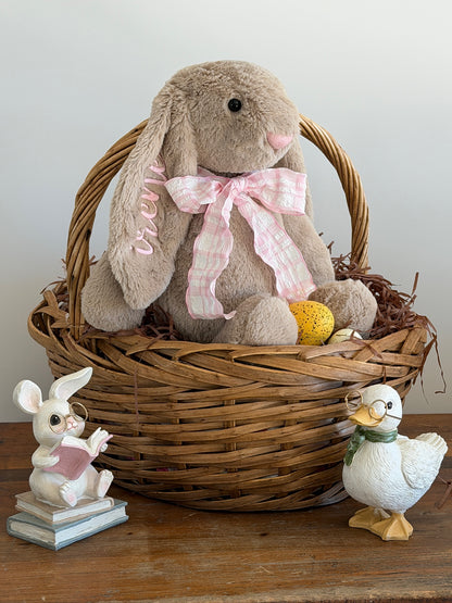 Stuffed bunny with a pink bow in a wicker basket with small figurines on a wooden surface.