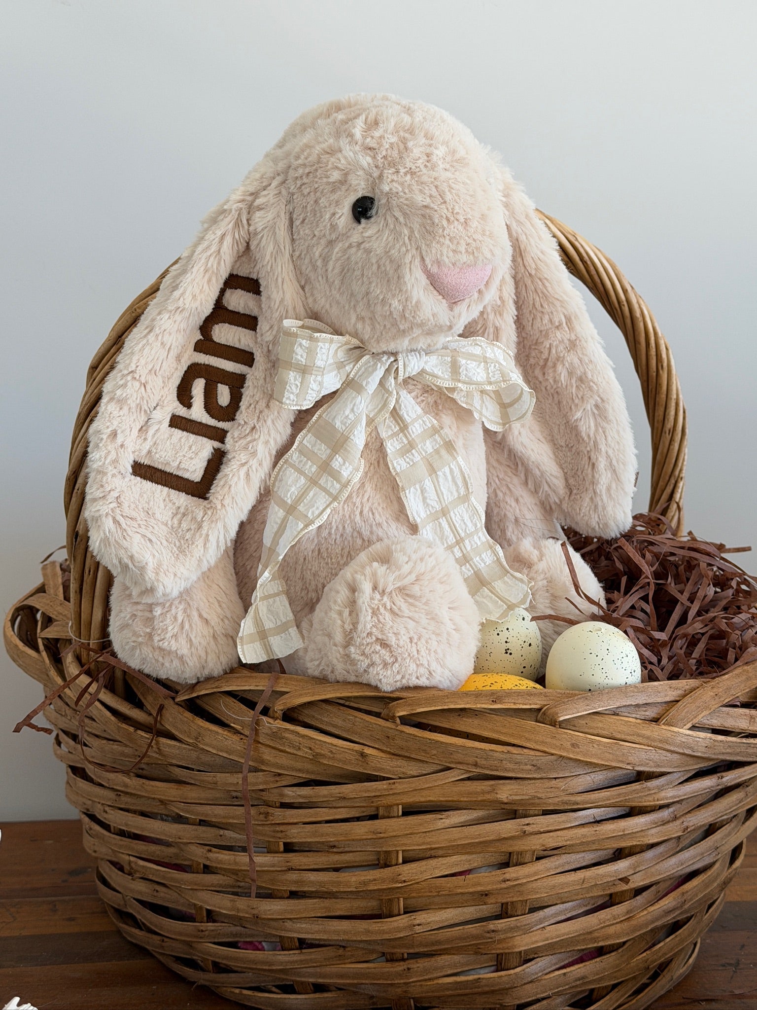 Plush bunny toy with a bow named 'Liam' in a wicker basket with Easter eggs and grass.