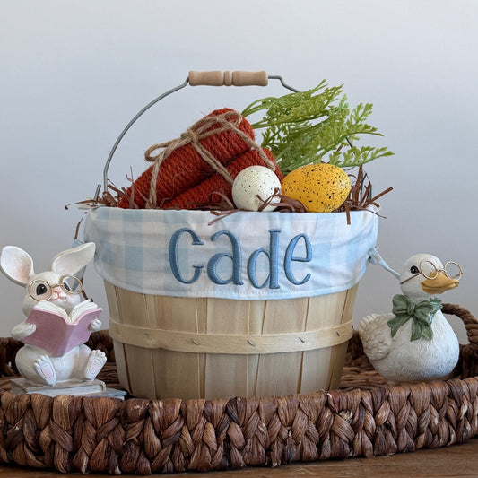 Decorative basket with embroidered name 'Cade', surrounded by small figurines and plants on a wooden surface.