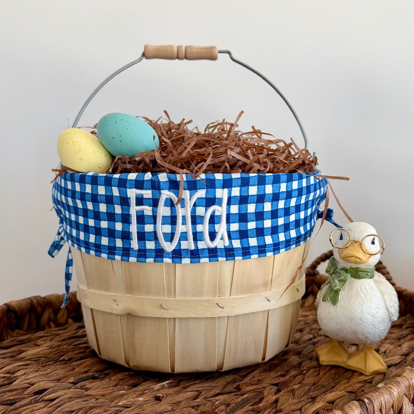 Wooden Easter basket with blue checkered ribbon, eggs, and a duck decoration on a woven surface.