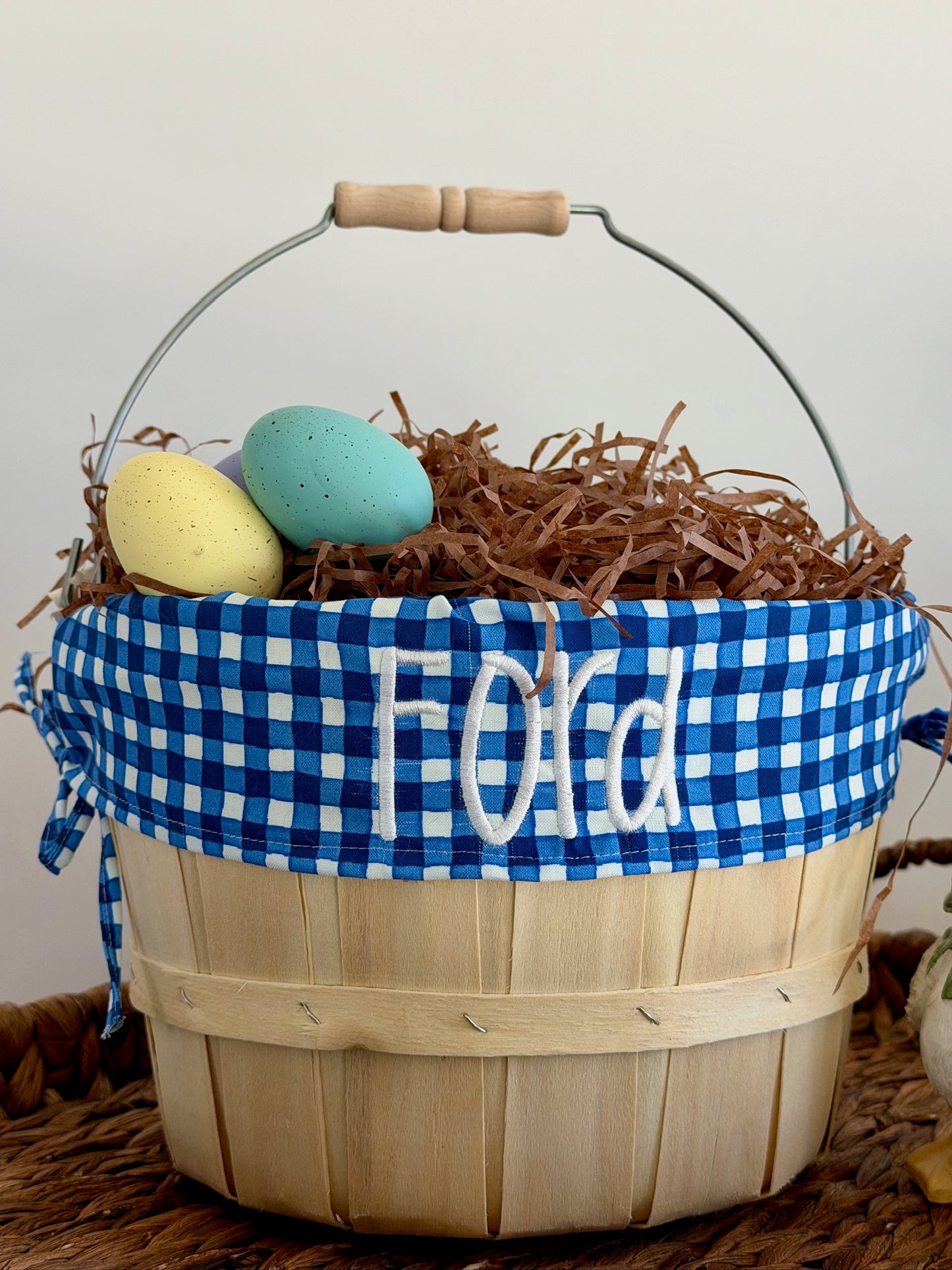 Wooden basket with blue checkered ribbon and embroidered name, filled with decorative eggs on a neutral background.