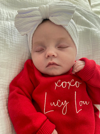 Newborn baby wearing a red outfit with 'XOXO Lucy Lou' text, lying on a textured white surface.