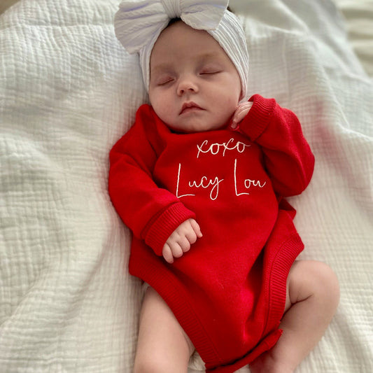 Newborn baby wearing a red outfit with 'xoxo Lucy Lou' text and a white headband on a white blanket.