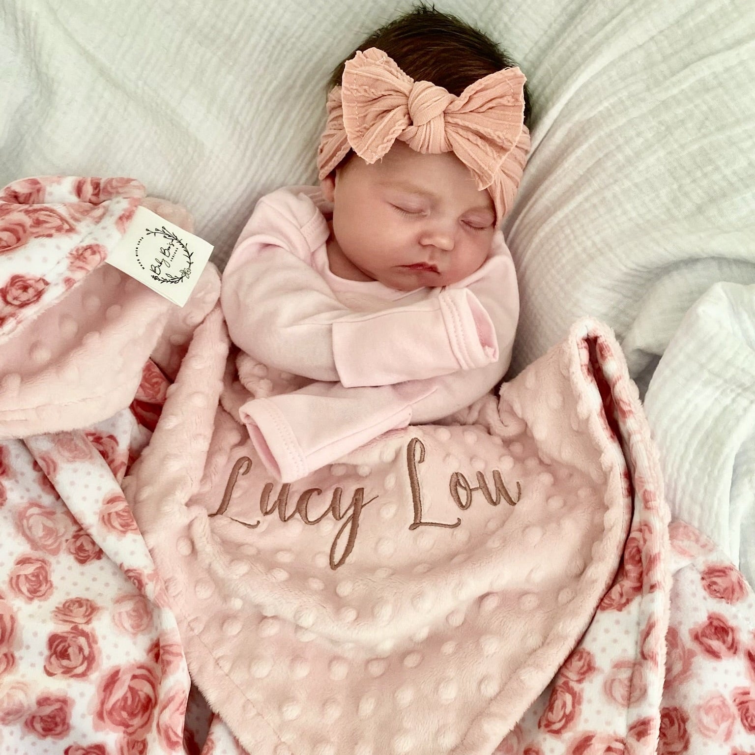 Newborn baby wrapped in a pink blanket with 'Lucy Lou' embroidered, wearing a matching headband, on a soft surface.