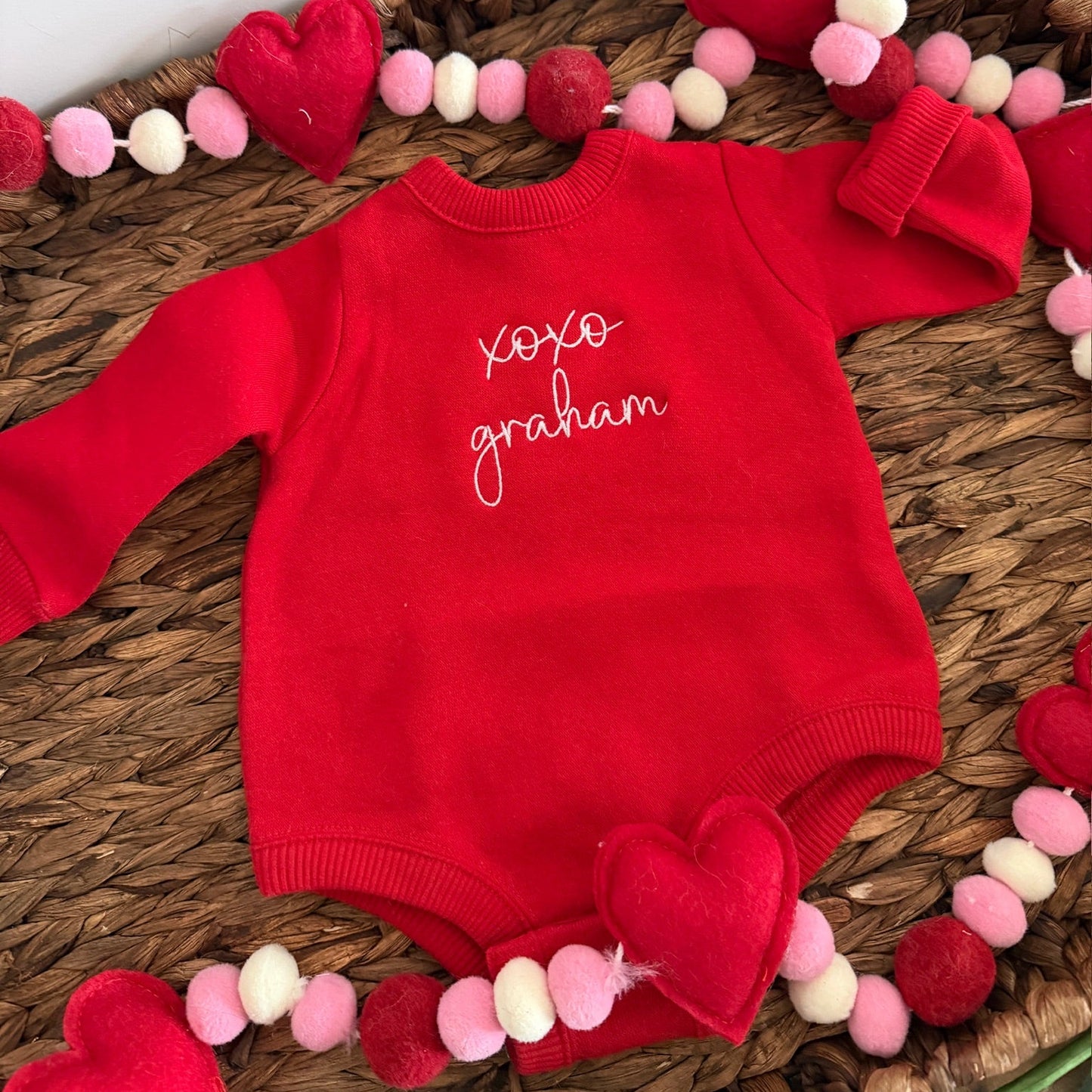 Red baby onesie with embroidered name on a decorative surface with heart-shaped ornaments