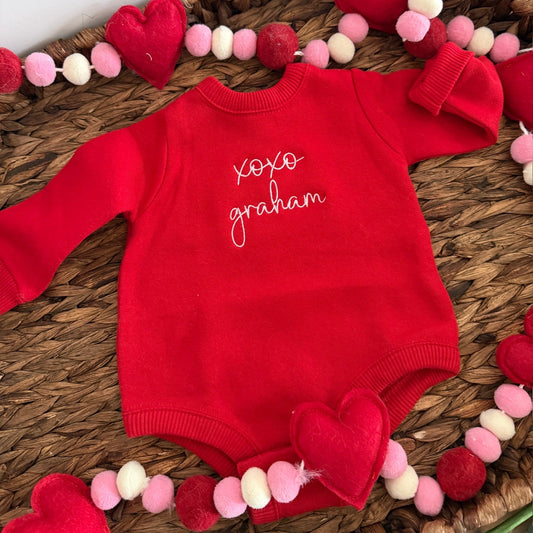 Red baby onesie with embroidered name on a decorative surface with heart-shaped ornaments
